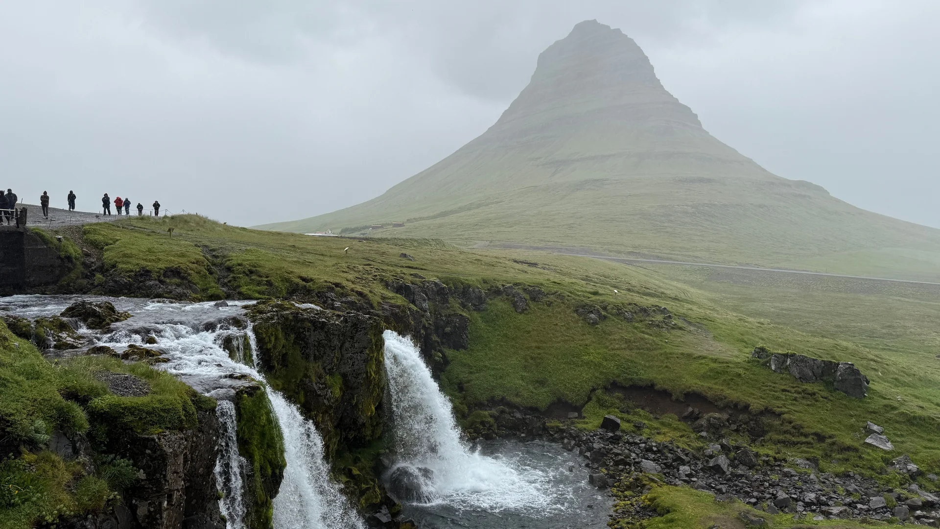 Kirkjufell mountain rising behind Kirkjufellsfoss waterfall on the Snaefellsnes Peninsula, with visitors watching from the cliff edge